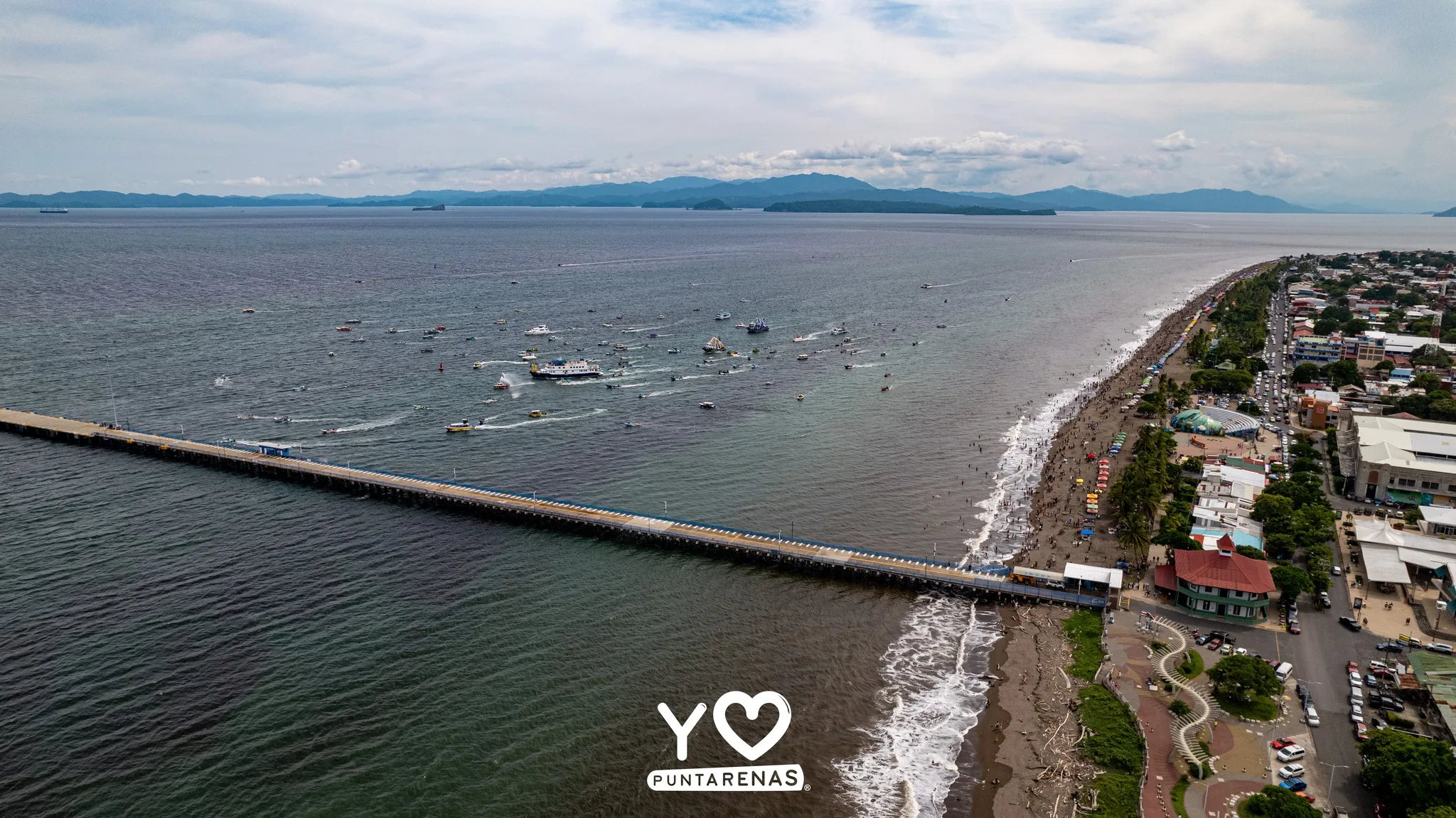 Vista aérea de Puntarenas durante la procesión marítima de la Virgen del Mar, con embarcaciones frente al muelle y la playa.